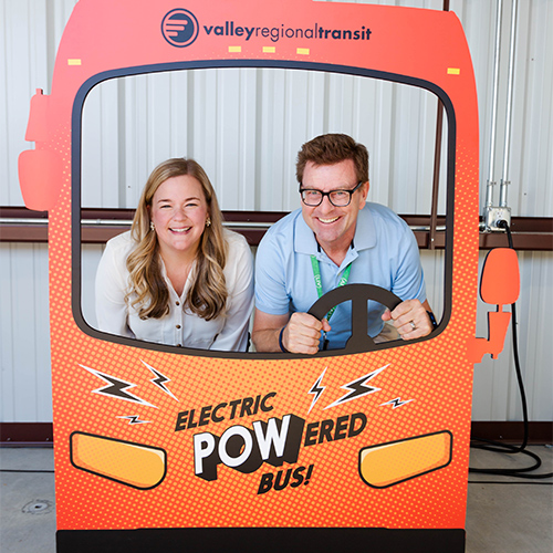 Two people sitting behind a cut out of an electric cardboard bus.