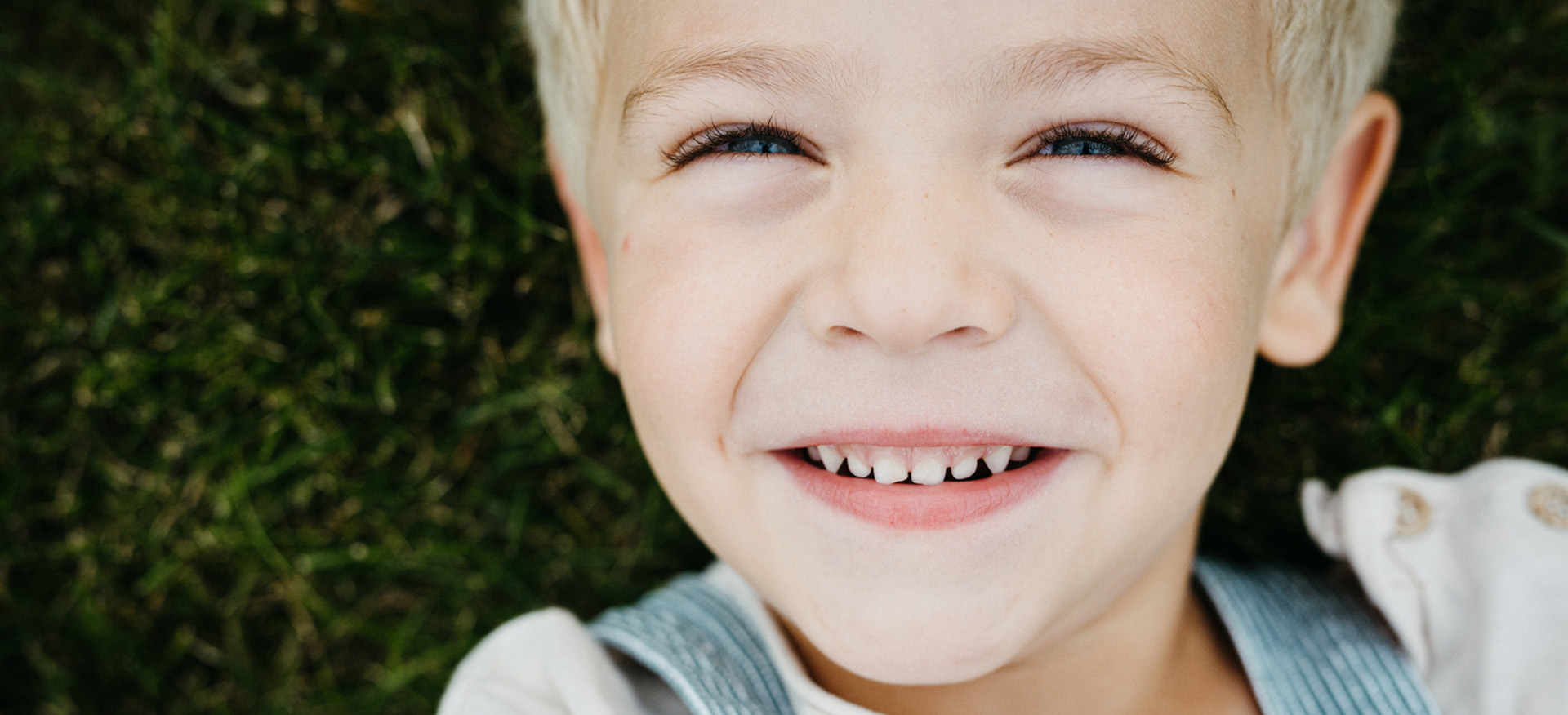 An overhead close up of a young boy with a smile on his face laying in the grass.