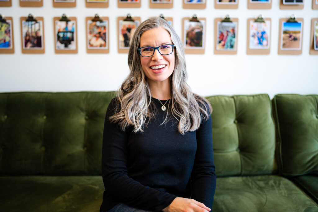 Photograph of a smiling woman, with gray hair, wearing glasses and a black shirt sitting on a green velvet couch in office.