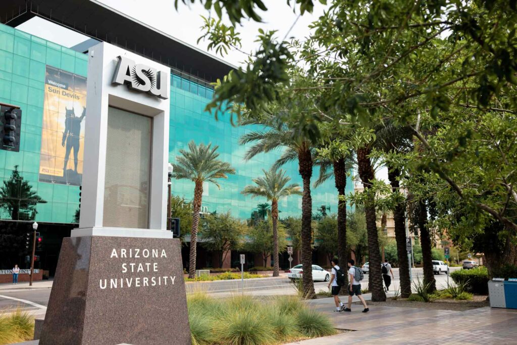 Arizona State University campus with the sign out front. Three students walking towards the building. 